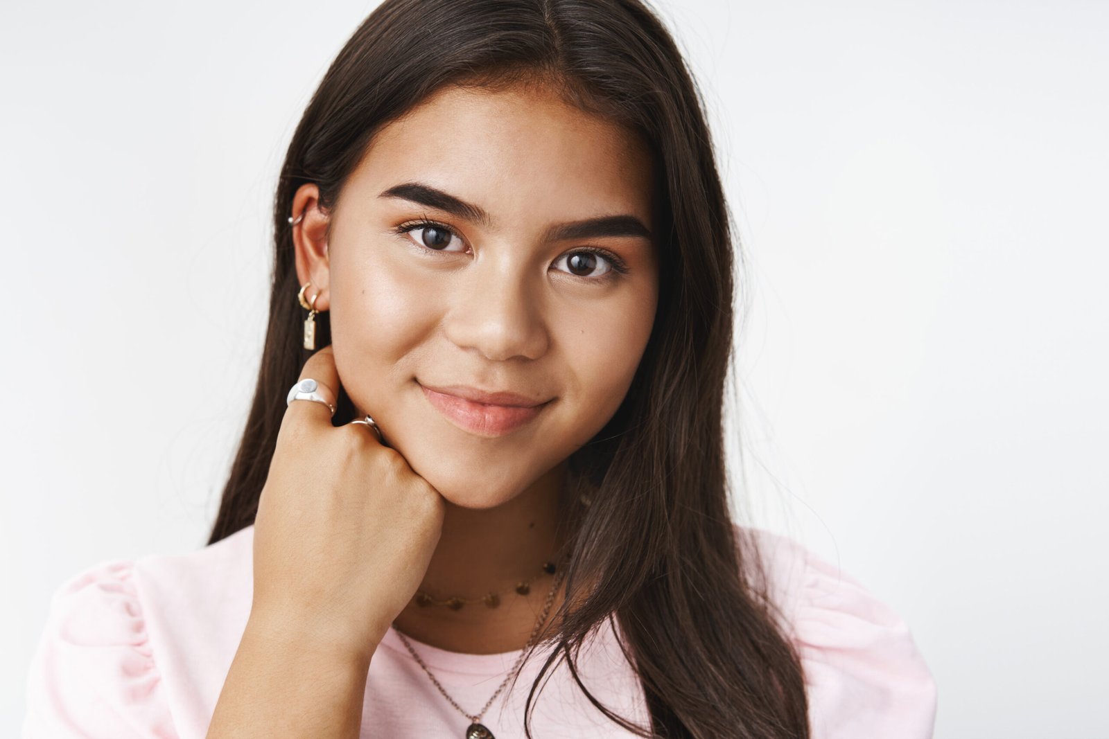Waist-up shot of cute and kind sincere indian teenage girl with charming smile gazing at camera touching face with hand posing against gray background relaxed and carefree